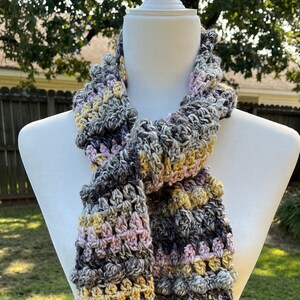 May include: A crocheted scarf in shades of gray, pink, and yellow. The scarf is made with a textured stitch and has a fringed edge.