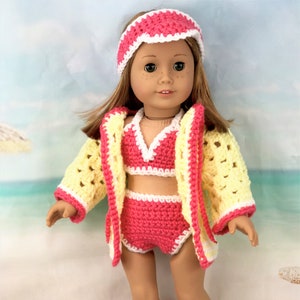 Op de afbeelding: Een roze en gele gehaakte badpak en badjas set voor een pop. De set bevat een hoofdband, een bikini top, een rok en een badjas. De pop draagt de set en staat op een strand.