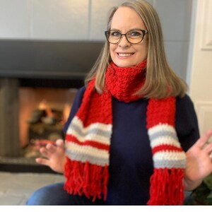 May include: A red and white striped scarf with fringe. The scarf is crocheted and has a textured pattern. The scarf is worn by a person with long brown hair and glasses.