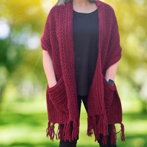 May include: A burgundy crochet shawl with fringe and two pockets. The shawl is worn over a black shirt and black pants.