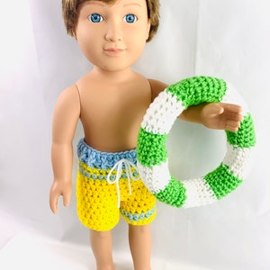 Könnte beinhalten: Eine Puppe trägt eine gelbe, blaue und grüne gehäkelte Badehose und hält einen grünen und weißen gehäkelten Schwimmreifen.