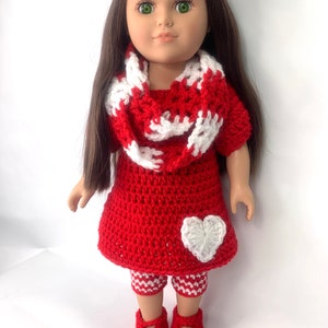 May include: A red crocheted dress with a white heart detail, red and white crocheted leggings, and a red and white crocheted scarf. The doll is wearing red crocheted shoes.