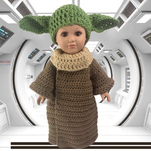 Puede incluir: Una muñeca con una túnica de ganchillo marrón y un gorro de ganchillo verde con orejas. La muñeca está de pie en un pasillo blanco con un cartel de "WELCOME" en la pared.