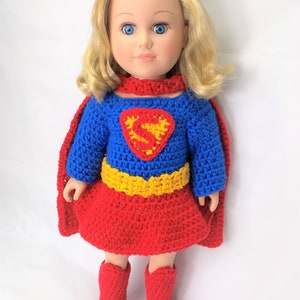 May include: A doll with blonde hair and blue eyes wearing a handmade crochet superhero costume. The costume includes a blue top with a red and yellow emblem, a red skirt, cape, and boots. The doll is standing against a white background.