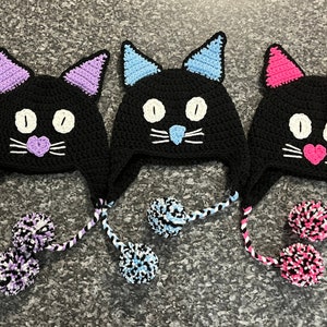 May include: Three black crocheted cat hats with pom-poms. The hats have white eyes, a pink nose, and a white mustache. The pom-poms are black, white, and pink.