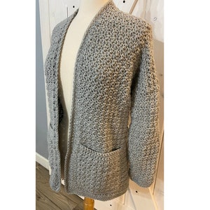 May include: A gray crocheted cardigan with long sleeves and two pockets. The cardigan is open in the front and has a simple, classic design.