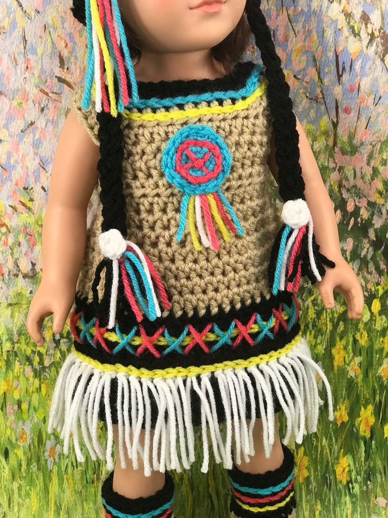 May include: A crocheted doll outfit in brown, black, white, yellow, pink, and blue. The outfit includes a dress with fringe, a hat, and leg warmers. The dress has a beaded design and a dreamcatcher motif.
