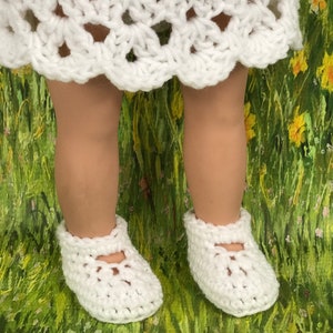 May include: A pair of white crochet baby shoes with a Mary Jane style strap. The shoes are shown on a doll's feet, with a white crochet dress visible above. The background is a green and yellow floral pattern.