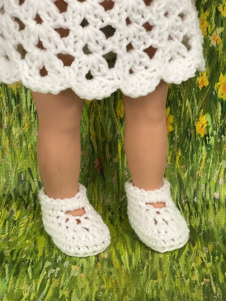 May include: A pair of white crochet baby shoes with a Mary Jane style strap. The shoes are shown on a doll's feet, with a white crochet dress visible above. The background is a green and yellow floral pattern.