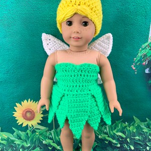 May include: A green and yellow crocheted Tinkerbell costume for a doll. The costume includes a dress, hat, and shoes. The doll is wearing the costume and has a white crocheted halo.