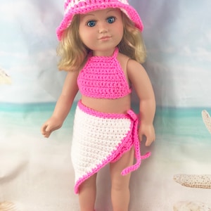 May include: A pink and white crocheted swimsuit and hat for a doll. The swimsuit has a halter top and a wrap skirt. The doll is wearing pink crocheted shoes.