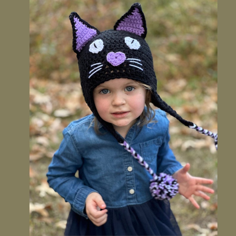 May include: A child wearing a black crocheted cat hat with purple ears and a white pom-pom on the end of the tie. The hat has white whiskers and a purple heart nose.