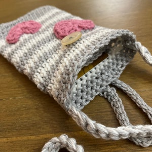 May include: A small grey and white crocheted bag with a pink heart design and a white button. The bag has a long strap and is ideal for carrying small items.