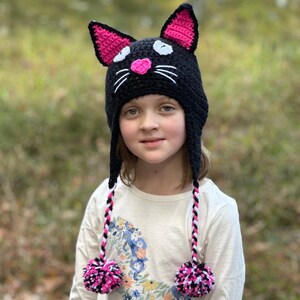 May include: A black crocheted cat hat with pink ears and a white pom-pom on each end of the long ties. The hat has white and black stripes on the ties and a pink nose.