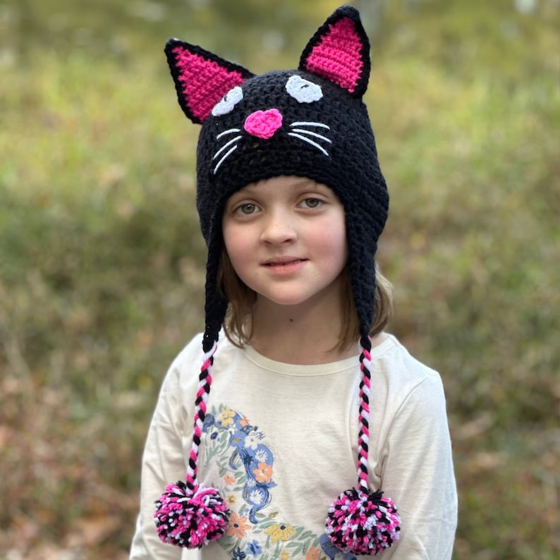 May include: A black crocheted cat hat with pink ears and a white pom-pom on each end of the long ties. The hat has white and black stripes on the ties and a pink nose.