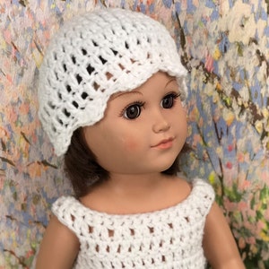 May include: A white crochet hat and dress on a doll. The hat has a scalloped edge, and the dress has a sleeveless design. The doll has brown hair and is set against a colorful floral background.