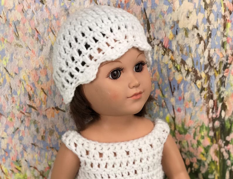 May include: A white crochet hat and dress on a doll. The hat has a scalloped edge, and the dress has a sleeveless design. The doll has brown hair and is set against a colorful floral background.