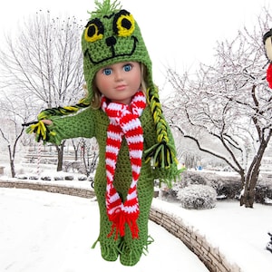 Op de afbeelding: Een groene gehaakte poppenoutfit met een Grinch-thema hoed, sjaal en bijpassende broek. De pop draagt een rood-wit gestreepte sjaal. De pop staat in een besneeuwde omgeving.