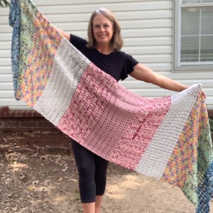 May include: A woman holds a long, colorful crocheted scarf with different colored stripes. The scarf is made of a variety of yarns in shades of blue, green, pink, white, and yellow.