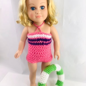 Könnte beinhalten: Eine Puppe trägt einen pinkfarbenen, lila und weißen gestreiften gehäkelten Badeanzug und hält einen grün-weiß gestreiften gehäkelten Schwimmreifen.
