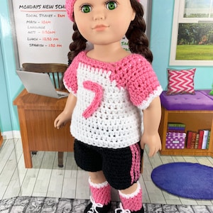 May include: A doll wearing a pink and white crocheted jersey with the number 7 on the front, black shorts with pink stripes, and pink and white crocheted socks with black shoes.