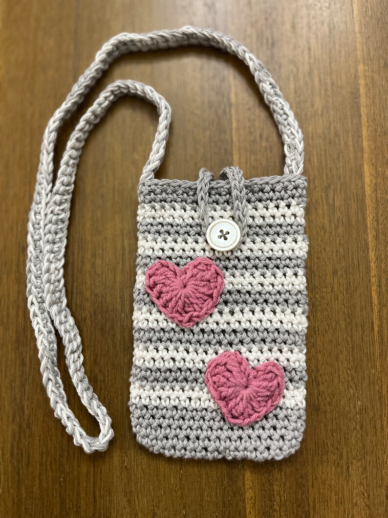 May include: A grey and white striped crocheted crossbody bag with a pink heart design and a white button closure.