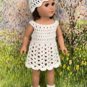 May include: A white crochet doll outfit, including a dress, hat, and shoes. The dress has a scalloped hem and openwork design. The hat has a scalloped brim. The shoes are Mary Jane style. The doll is standing in front of a colorful background.