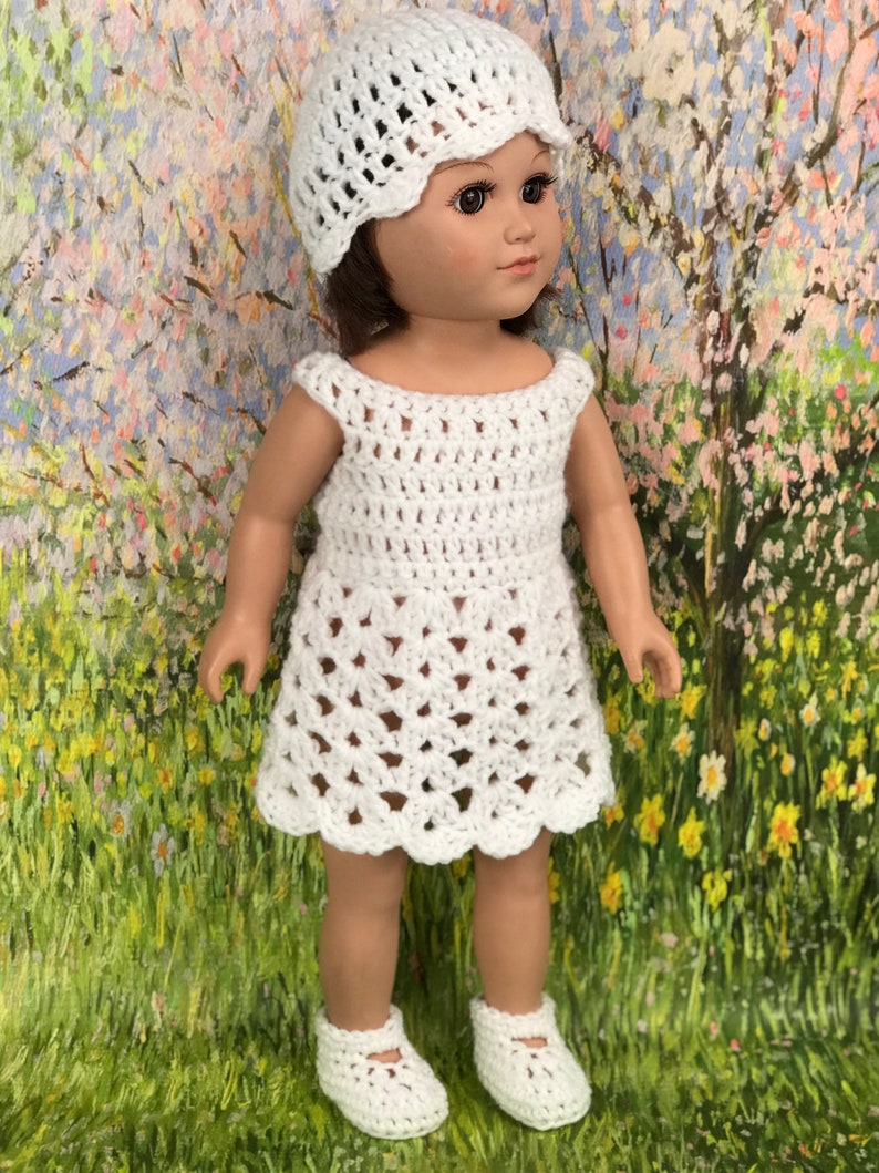 May include: A white crochet doll outfit, including a dress, hat, and shoes. The dress has a scalloped hem and openwork design. The hat has a scalloped brim. The shoes are Mary Jane style. The doll is standing in front of a colorful background.