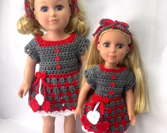 May include: Two dolls wearing matching gray and red crochet dresses with white heart accents. The dolls have blonde hair and blue eyes. They are wearing red shoes.