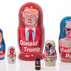 Könnte beinhalten: Satz von sechs Nistpuppen, oder Matroschka-Puppen, mit politischen Figuren. Die größte Puppe ist rot und zeigt Donald Trump. Die nächstgrößte Puppe ist blau und zeigt Wladimir Putin. Die drittgrößte Puppe ist blau und zeigt Hillary Clinton. Die viertgrößte Puppe ist beige und zeigt eine Frau mit einem Schwert und einem Drachen. Die fünftgrößte Puppe ist rot und zeigt einen Footballspieler. Die kleinste Puppe ist schwarz und zeigt eine Sonnenfinsternis.