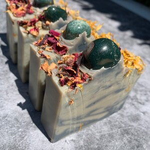 May include: Four bars of handmade soap with green gemstone accents and dried rose petals. The soap is a light gray with dark gray swirls.