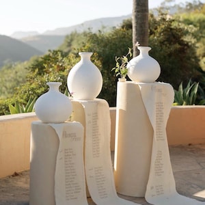 Puede incluir: Tres jarrones de cerámica blanca se asientan sobre pedestales cilíndricos, cada uno con un rollo de papel que enumera nombres. Los jarrones tienen forma redondeada con cuello estrecho. Los rollos de papel son de color blanquecino y presentan texto manuscrito. La escena es al aire libre.