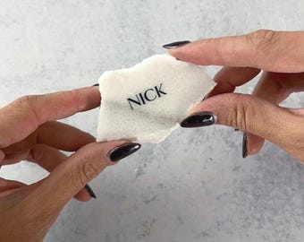May include: A small, irregularly shaped white stone with the name "NICK" printed in black. The stone is held between two hands against a light gray background. The hands have dark nail polish.