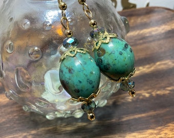 May include: A pair of dangle earrings with green and gold accents. The earrings feature a large, oval-shaped green bead with gold detailing and a smaller, clear crystal bead hanging below.