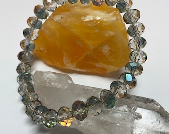 May include: A crystal bracelet with iridescent, faceted beads in shades of gray and green. The bracelet is displayed on a white and orange stone background.