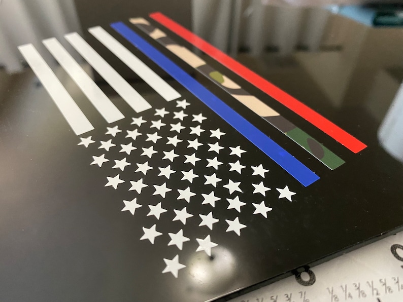 Puede incluir: Un fondo negro con un dise&ntilde;o de bandera estadounidense blanca. La bandera tiene una l&iacute;nea azul en el medio, una franja roja sobre la l&iacute;nea azul y un patr&oacute;n de camuflaje sobre la franja roja.