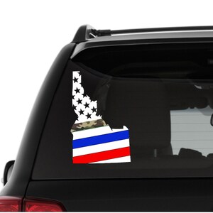 Może przedstawiać: Naklejka na szybę samochodu przedstawiająca zarys stanu Idaho, z wzorem kamuflażu, niebieskimi i czerwonymi paskami oraz białym tłem z gwiazdami.