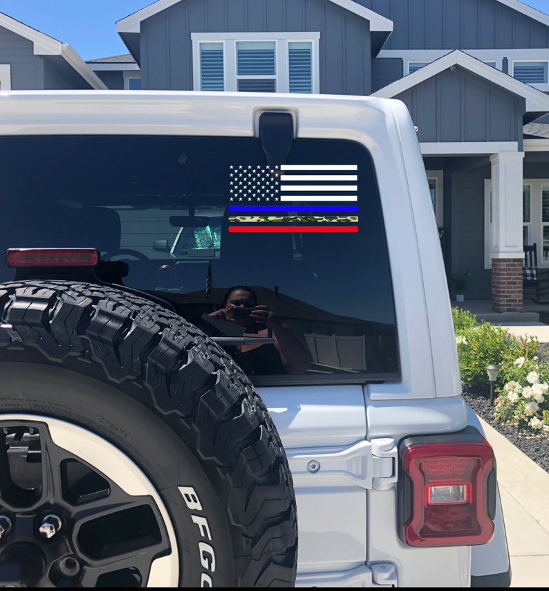 Puede incluir: Un Jeep blanco con una calcoman&iacute;a de bandera estadounidense de l&iacute;nea azul delgada en la ventana trasera. La calcoman&iacute;a tiene una l&iacute;nea roja delgada, una l&iacute;nea amarilla delgada y un patr&oacute;n de camuflaje.