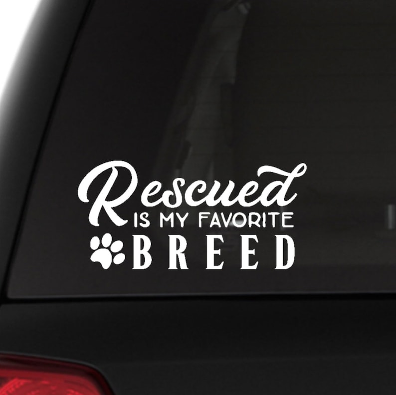 Puede incluir: Calcoman&iacute;a de vinilo blanco con el texto "Rescued is my favorite breed" y una huella de pata. La calcoman&iacute;a est&aacute; en una ventana de coche negro.