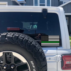 Puede incluir: Un Jeep blanco con una pegatina de bandera estadounidense negra y verde en la ventana trasera. La bandera tiene una fina franja verde en la parte inferior.