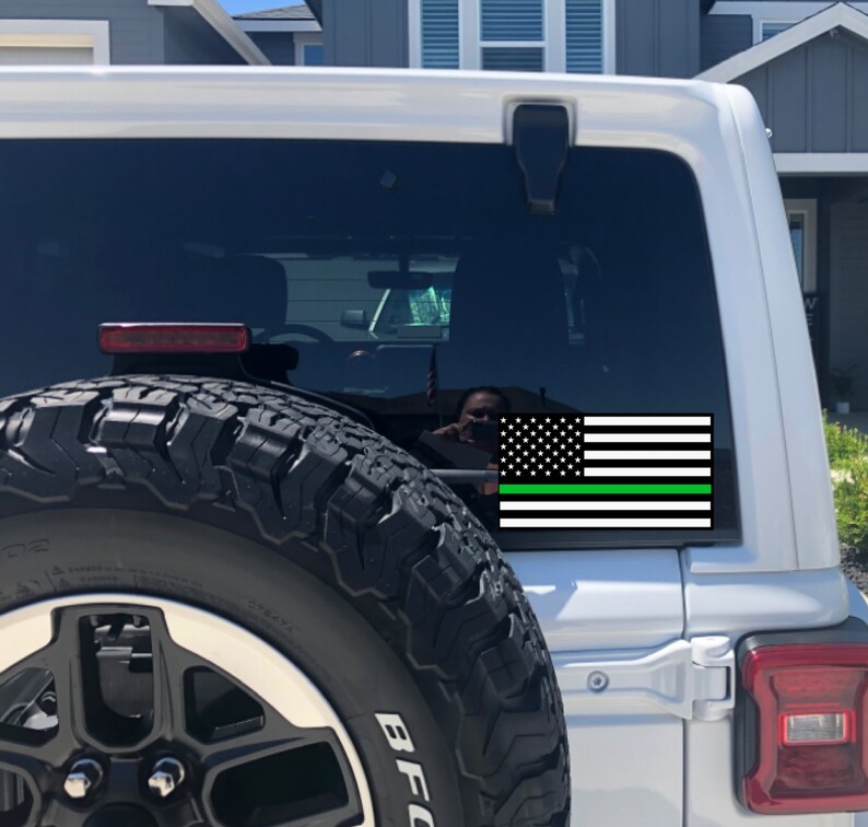 Puede incluir: Un Jeep blanco con una pegatina de bandera estadounidense negra y verde en la ventana trasera. La bandera tiene una fina franja verde en la parte inferior.
