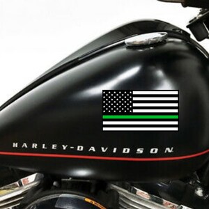 Puede incluir: Dep&oacute;sito de gasolina de motocicleta negro con un logotipo de Harley-Davidson y una calcoman&iacute;a de bandera estadounidense con una fina l&iacute;nea verde.