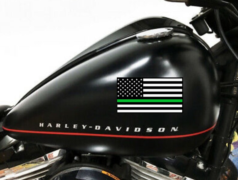 Puede incluir: Dep&oacute;sito de gasolina de motocicleta negro con un logotipo de Harley-Davidson y una calcoman&iacute;a de bandera estadounidense con una fina l&iacute;nea verde.