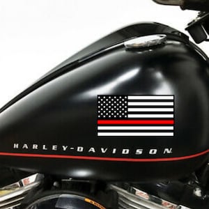 May include: Black motorcycle gas tank with a red, white, and blue American flag decal with a red stripe across the bottom. The tank has the words "Harley-Davidson" printed on it.