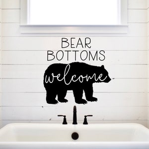May include: A black bear silhouette with the text "BEAR BOTTOMS welcome" on a white bathroom wall with a white bathtub.