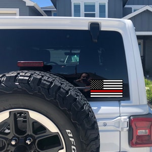 May include: A white Jeep with a black and red American flag sticker on the back window. The flag has a thin red stripe across the bottom, representing the thin red line of firefighters.