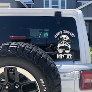 Puede incluir: Un Jeep blanco con una llanta negra y una pegatina negra y amarilla en la ventana trasera. La pegatina dice "The Voice of Courage & Hope Dispatcher".