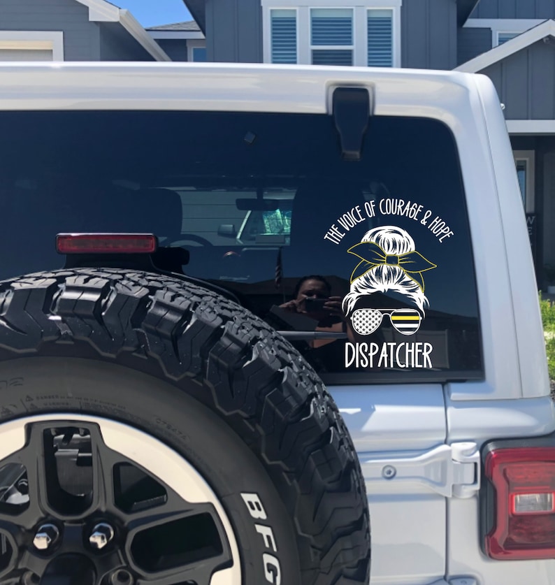 Puede incluir: Un Jeep blanco con una llanta negra y una pegatina negra y amarilla en la ventana trasera. La pegatina dice "The Voice of Courage & Hope Dispatcher".