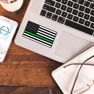 Puede incluir: Una pegatina de bandera estadounidense con una fina l&iacute;nea verde en un ordenador port&aacute;til. La pegatina es negra y blanca con una fina l&iacute;nea verde en el medio.