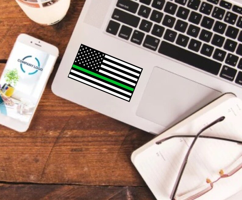 Puede incluir: Una pegatina de bandera estadounidense con una fina l&iacute;nea verde en un ordenador port&aacute;til. La pegatina es negra y blanca con una fina l&iacute;nea verde en el medio.